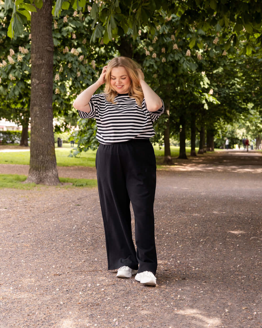 Nainen seisoo puistossa päällään Toiveen basics-malliston Noki housut ja raidallinen Muisto t-paita.