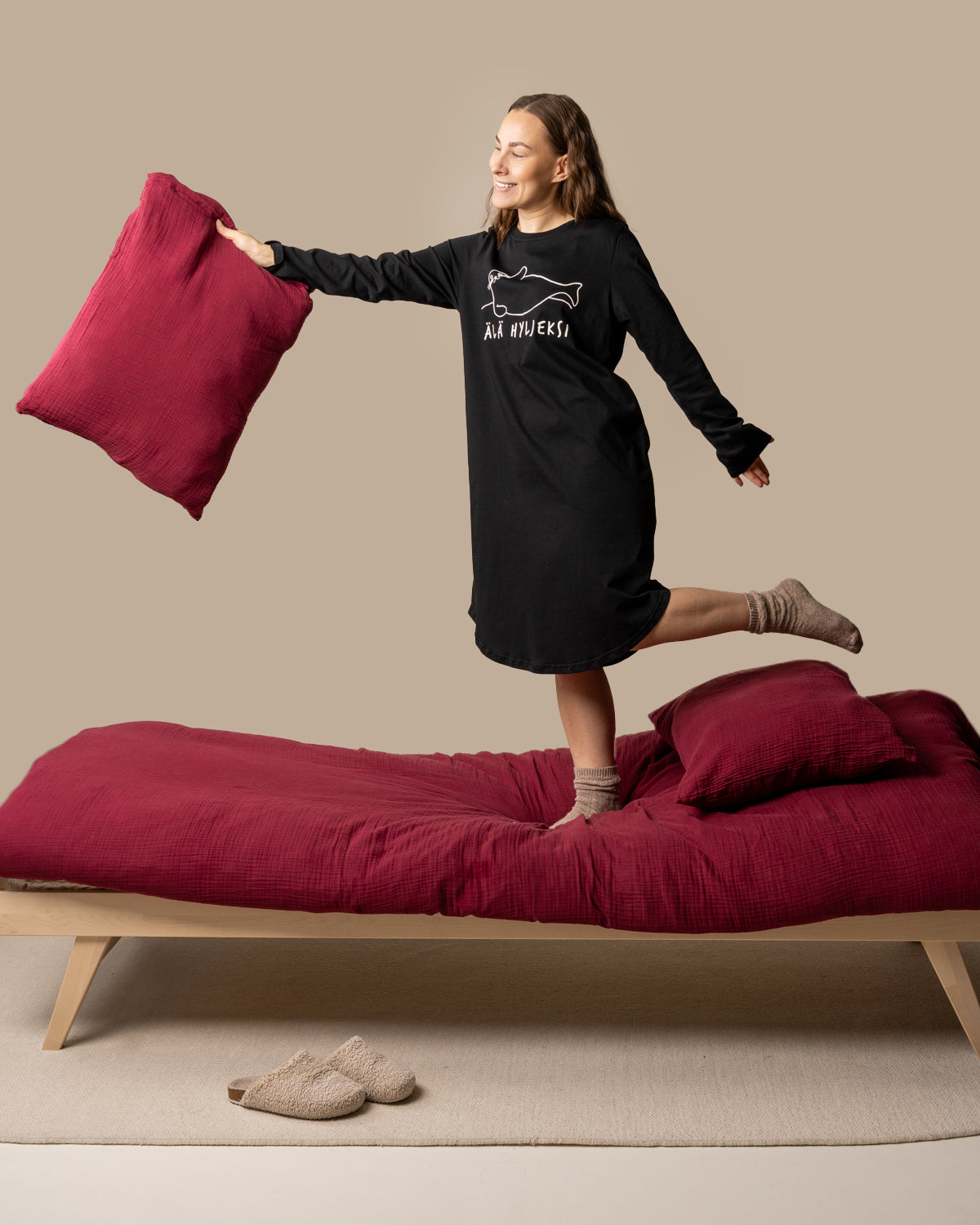 Nainen mustassa Älä hyljeksi yömekossa heittää tyynyä, taustalla sänky ja punaiset tyynyt.